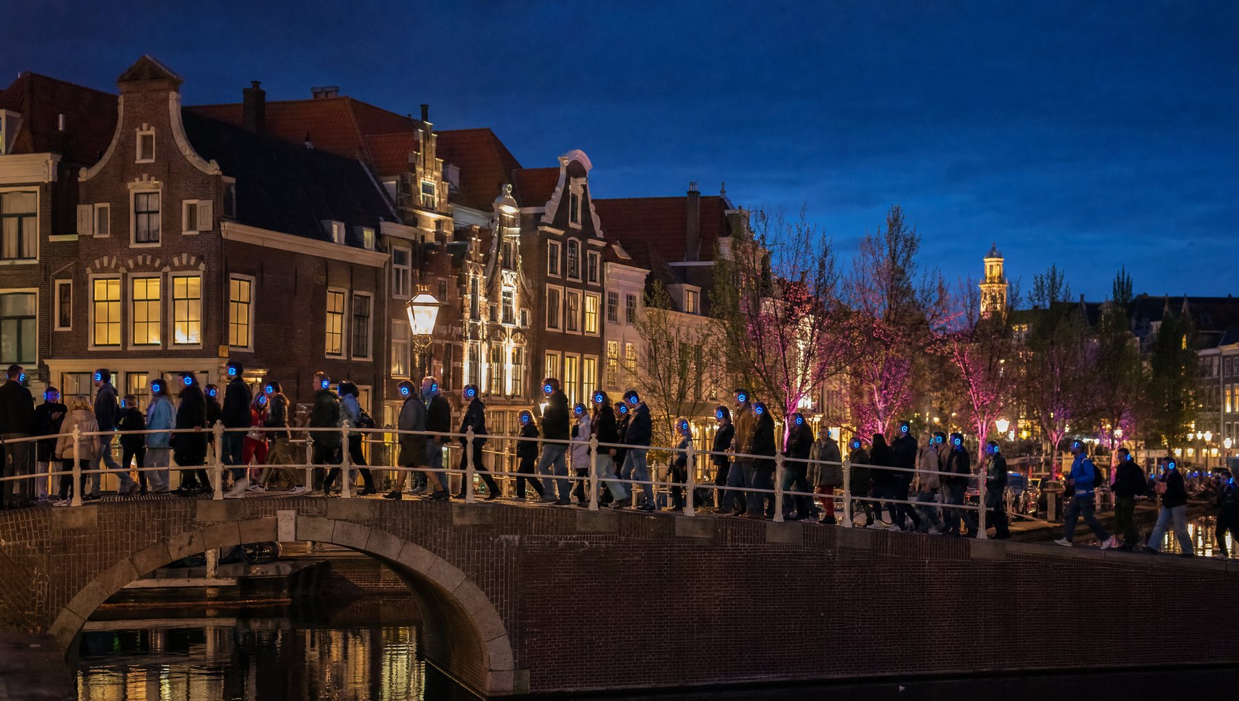 Sfeerimpressie: een avondwandeling over een brug langs de gracht bij schemer, iedereen met een gloeiende koptelefoon op